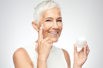 Gorgeous smiling Caucasian senior woman trying out new anti age cream and looking at camera. Beauty...
