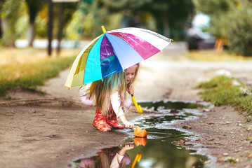 Girl with colorful umbrella making paper ship sail in puddle, happy childhood