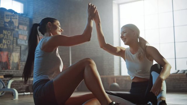 Two Beautiful Fit Athletic Girls Sit on a Floor of Industrial Loft Gym. They're Happy with their Training Program and Successfully Give a High Five. Strong Masculine Man Walks in the Background.