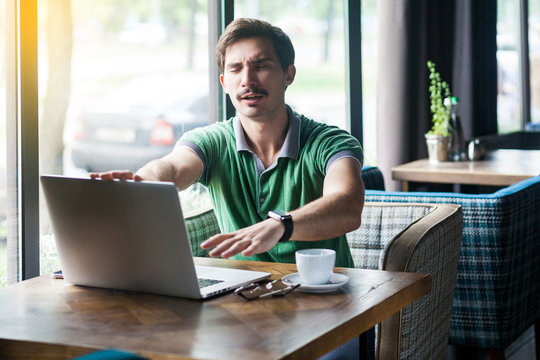 Blindness and way of business. Young blind businessman in green t-shirt sitting and trying to find his laptop with closed eyes. business and freelancing concept. indoor shot near big window at daytime