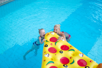 Beautiful family Mom and daughter swim in a hotel pool with blue water and eat a funny pizza air...