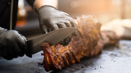 Grill restaurant kitchen. Chef in black cooking gloves using knife to cut smoked pork ribs. - obrazy, fototapety, plakaty