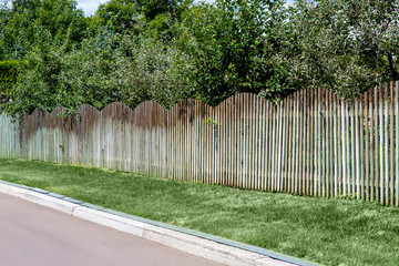 different fruit orchard behind a wooden fence at the side of the asphalt road with a curb and drainage system and green lawn.