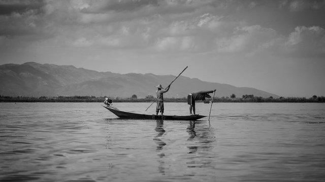 Sylwetki rybak&oacute;w z Inle Lake wiosłujących po jeziorze na tle g&oacute;r - czarno białe.