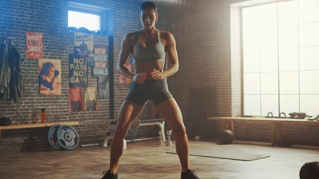 Strong and Fit Beautiful Athletic Woman in Sport Top and Shorts is Doing Squat Exercises in a Loft Style Industrial Gym with Motivational Posters