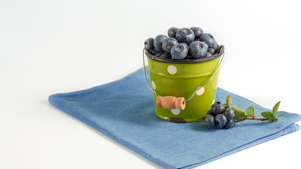 On a blue napkin on a white background is a small green bucket filled with blueberries. Healthy and tasty food, organic food. Selective focus. The concept of healthy eating. Horizontal. Copy space.