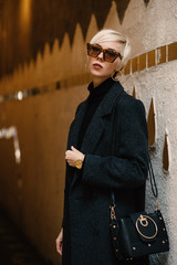 Street fashion portrait of young elegant blonde woman wearing autumn dark gray coat, sunglasses...