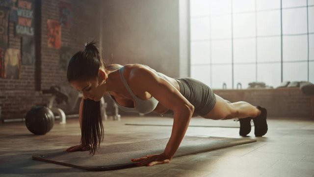Strong and Fit Athletic Woman in Sport Top and Shorts is Doing Push Up Exercises in a Loft Style Industrial Gym with Motivational Posters. It's Part of Her Cross Fitness Training Workout. Warm Light.