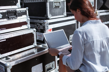 Installation of stage equipment and preparing for a live concert open air. Event manager portrait. Summer music city festival. Young serious woman stand and work with her laptop near the stage.