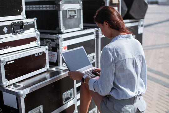Installation of stage equipment and preparing for a live concert open air. Event manager portrait. Summer music city festival. Young serious woman stand and work with her laptop near the stage.