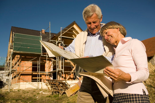 Senior couple looking at blueprints in front of incomplete built house, smiling