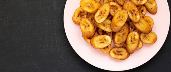 Homemade fried plantains on a pink plate on a black background, top view. Flat lay, from above, overhead. Copy space.