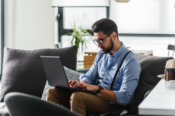 Young bearded businessman working on laptop computer while sitting on sofa at his home office. Business problems.