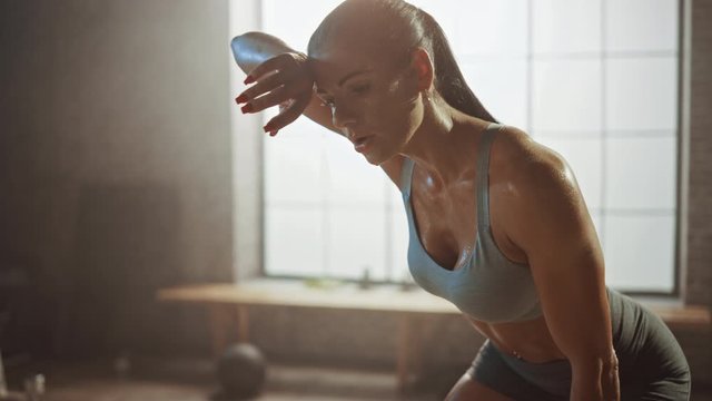 Portrait of a Beautiful Strong Fit Brunette Wiping Sweat from Her Face in a Loft Industrial Gym with Motivational Posters. She's Catching Her Breath after Intense Fitness Training Workout. Warm Light.