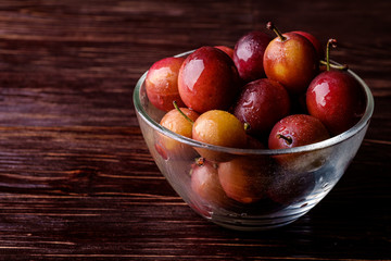 Ripe sweet plum fruits with water drops in glass bowl on dark moody wood table background