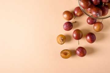 Scattered sliced half ripe sweet plum fruits with water drops near to plums in glass bowl on cream colored background, bright, top view, flat lay