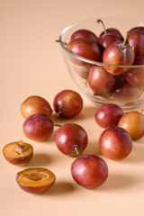 Scattered sliced half ripe sweet plum fruits with water drops near to plums in glass bowl on cream colored background, bright, angle view, selective focus