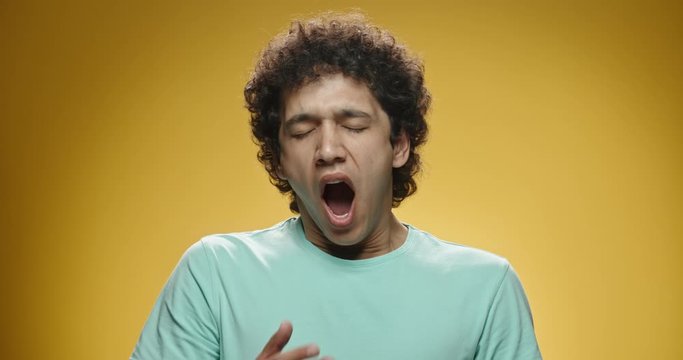 Young asian man with curly hair in casual clothes beind tired or bored, yawning and covering his mouth, isolated on yellow background close up 4k