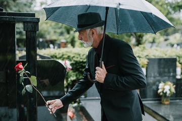 Elegant sad elderly man standing on the rain with umbrella and grieves at the grave of a loved...