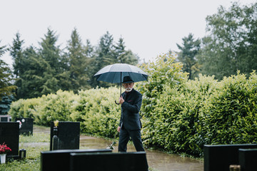 Elegant sad elderly man with black suite and umbrella walking alone on graveyard. Rainy day.