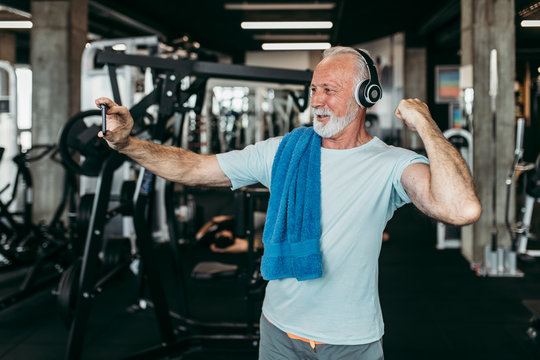 Senior man with headphones posing and taking selfie photo while exercising at gym.