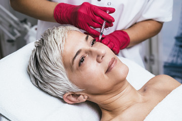 Fototapeta na wymiar Mature woman is getting a rejuvenating facial injections. She is lying calmly at clinic. The expert beautician is injecting botox into woman's wrinkles.