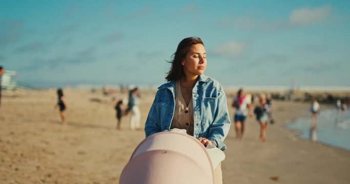 Young mother walks by the beach with her newborn baby, everyday family moment, watching other people, walk by seaside with buggy