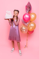 A teenage girl with balloons holds a cake in her hands, on a pink background