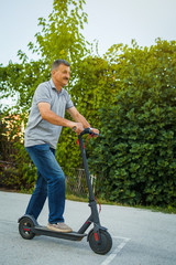 Senior man riding driving kick push scooter on the asphalt in front of the bushes green fence in a...