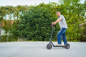 Senior man riding driving kick push scooter on the asphalt in front of the bushes green fence in a summer day