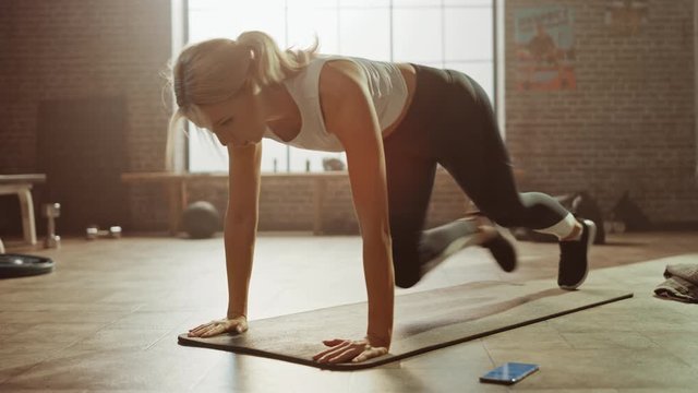 Beautiful and Young Girl Uses Smartphone App to Time Her Exercise and Starts doing Running Plank on Her Fitness Mat. Gorgeous and Athletic Woman Does Mountain Climber Workout in Stylish Hardcore Gym