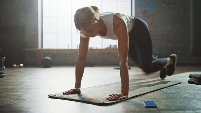 
Beautiful and Young Girl Uses Smartphone App to Setup Timer For Her Exercise and Starts doing Running Plank on Her Fitness Mat. Athletic Woman Does Mountain Climber Workout in Stylish Hardcore Gym
