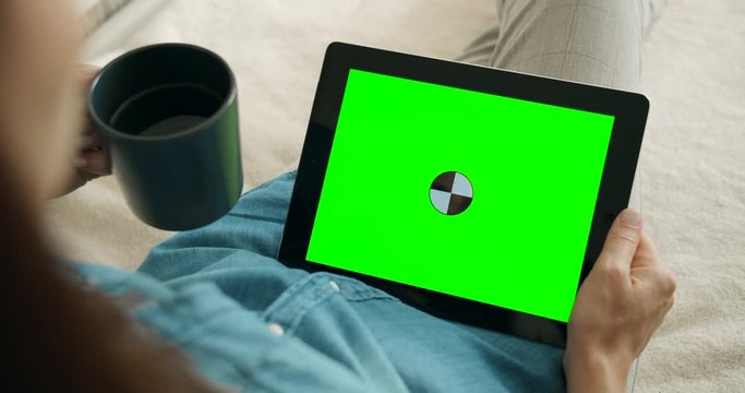 Woman resting on couch, holding greenscreen tablet and cup of coffee, advertisement template