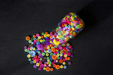 Top view of jar of buttons spilled out onto a black background; colorful buttons poured out of clear glass jar