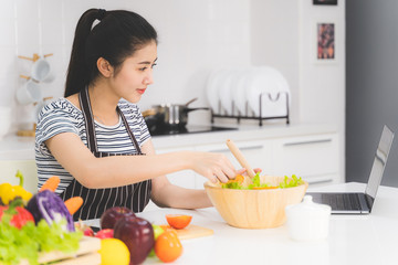 Beautiful young woman is learning to cook healthy food online by the internet from a laptop in white kitchen with many fresh vegetables and fruits on table.