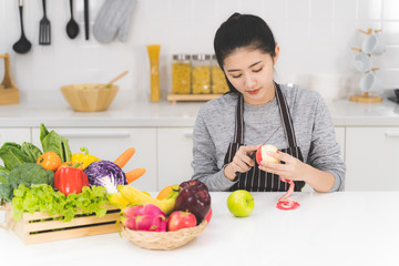 Beautiful asian woman, housewife peeling red apple in white kitchen with plenty of fresh vegetables and fruits on the table.
