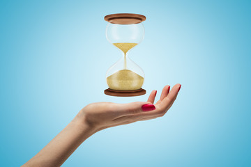 Side closeup of woman's hand facing up and levitating hourglass on light blue background.
