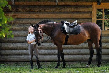 A teenage girl jockey stands next to a brown horse and hugs her. Against the background of a wooden wall stables.