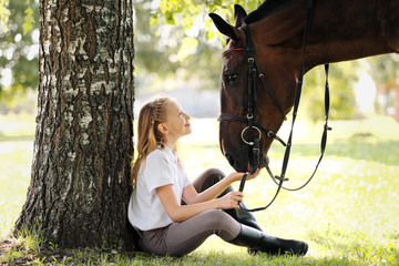 Girl teenager jockey sits in a green clearing under a tree. Feeds a horse an apple and strokes it.