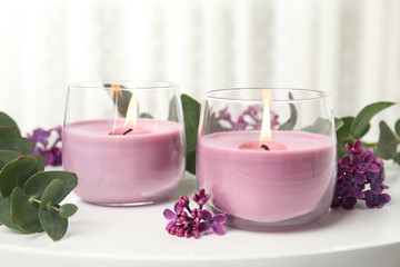 Fototapeta na wymiar Burning candles in glass holders and flowers with leaves on white table