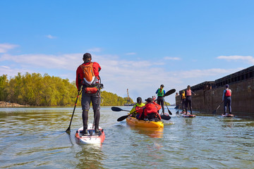 Peope on stand up paddle boards (SUP) and kayak paddling along the calm spring Danube river against a background of old rusty ships. Concept of water tourism, healthy lifestyle.