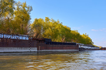 Old broken damaged rusty metal river barge on near the shore with green trees at early spring