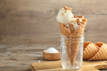 Waffle cone with ice cream, caramel and nuts in glass jar on wooden table. Space for text