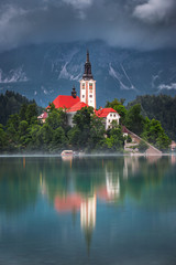 Fototapeta na wymiar Bled, Slovenia - The Pilgrimage Church of the Assumption of Maria surronded by fog on a small island and Julian Alps covered by clouds taken on a foggy summer morning at Lake Bled (Blejsko Jezero)