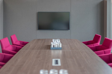 a brown conference table is surrounded by red armchairs and in the background there is a TV on the wall