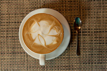 A white mug with a fragrant cappuccino with a pattern stands on a checkered litter. Near silver spoon.