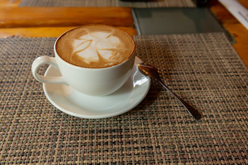 A white mug with a fragrant cappuccino with a pattern stands on a checkered litter. Near silver spoon.