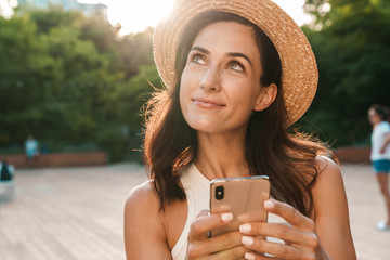 Image of dreaming adult woman typing on cellphone and looking upward at copyspace while walking in summer park