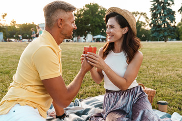 Image of happy middle-aged man giving present box to beautiful woman while sitting in park