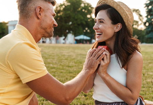 Image of smiling adult man giving present box to beautiful woman while sitting in park